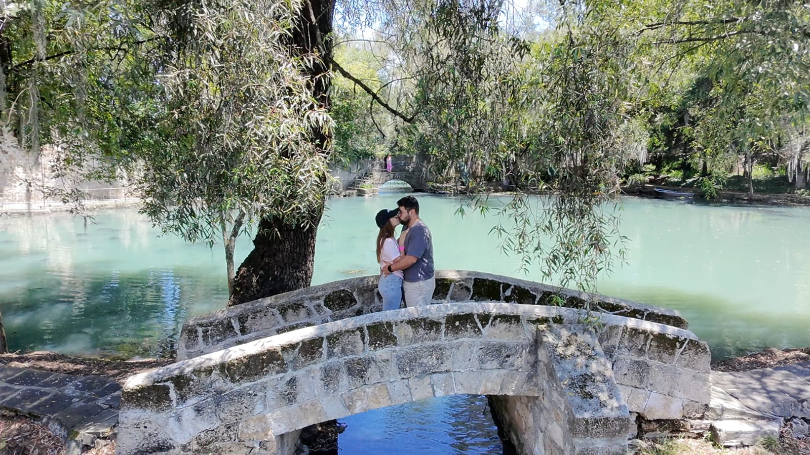 Alan y Lore besándose sobre un puente de piedra junto al agua